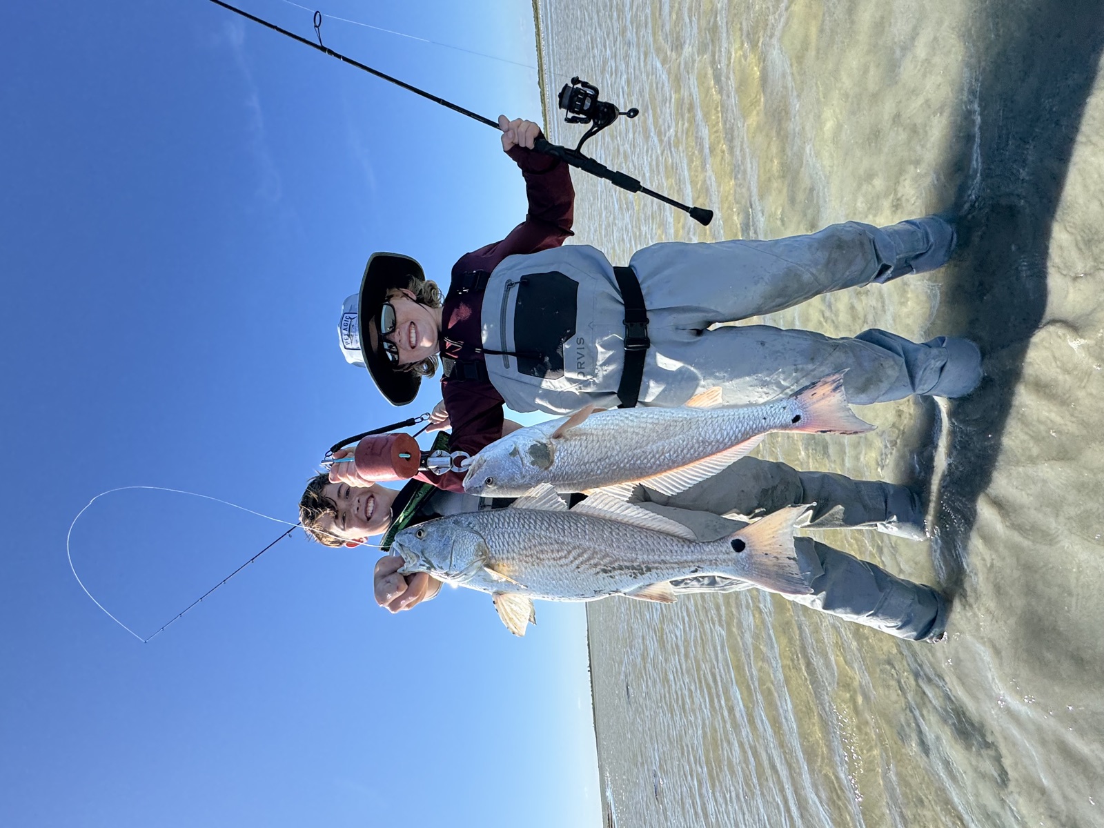 Two young anglers in waders holding up redfish on the flats